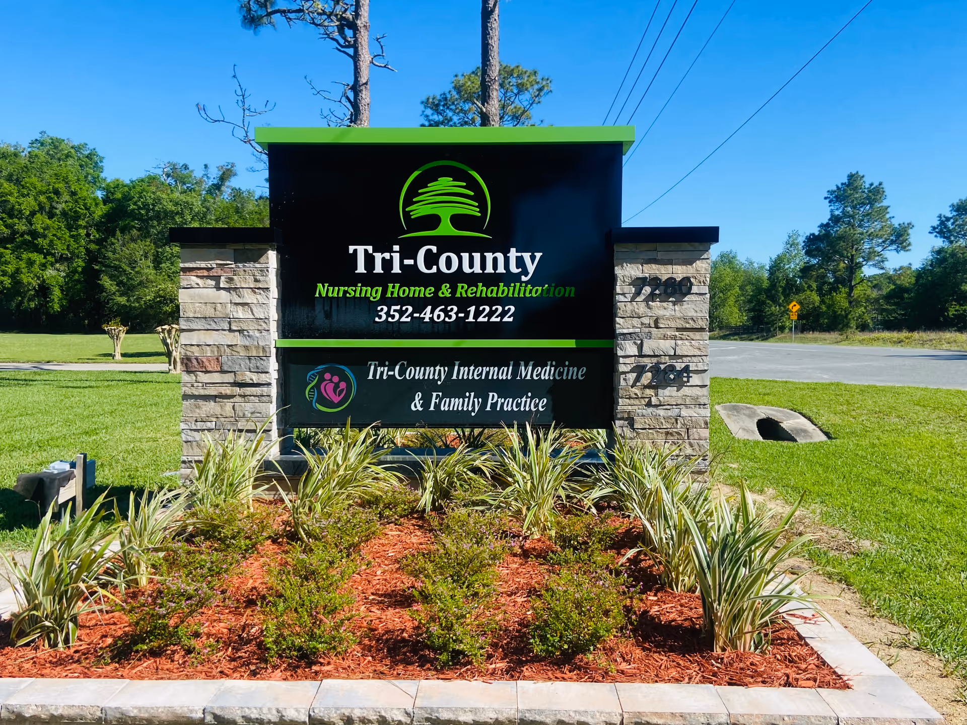 Outdoor sign for Tri-County Nursing Home & Rehabilitation and Tri-County Internal Medicine & Family Practice, surrounded by landscaped plants and grass with a clear blue sky in the background.