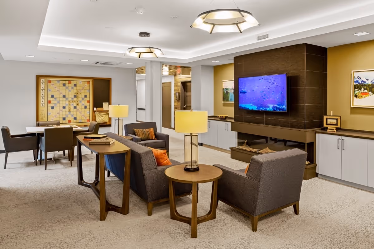 A cozy and modern living room area in a memory care facility featuring a gray sofa and two armchairs with orange accent pillows arranged around a round wooden side table with a lamp. A flat-screen TV is mounted on a dark wood-paneled wall above a built-in fireplace. In the background, there is a table with four chairs and a large Scrabble board on the wall. The room has soft lighting from ceiling fixtures and lamps, with neutral-colored walls and carpeted flooring.