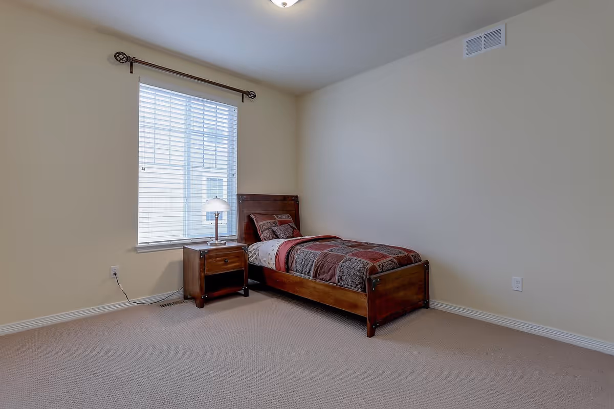 A simple bedroom with a single wooden bed featuring a patterned red and brown comforter and matching pillow. Next to the bed is a wooden nightstand with a lamp. A window with white blinds is on the left wall, and the room has beige walls and carpeted floor.