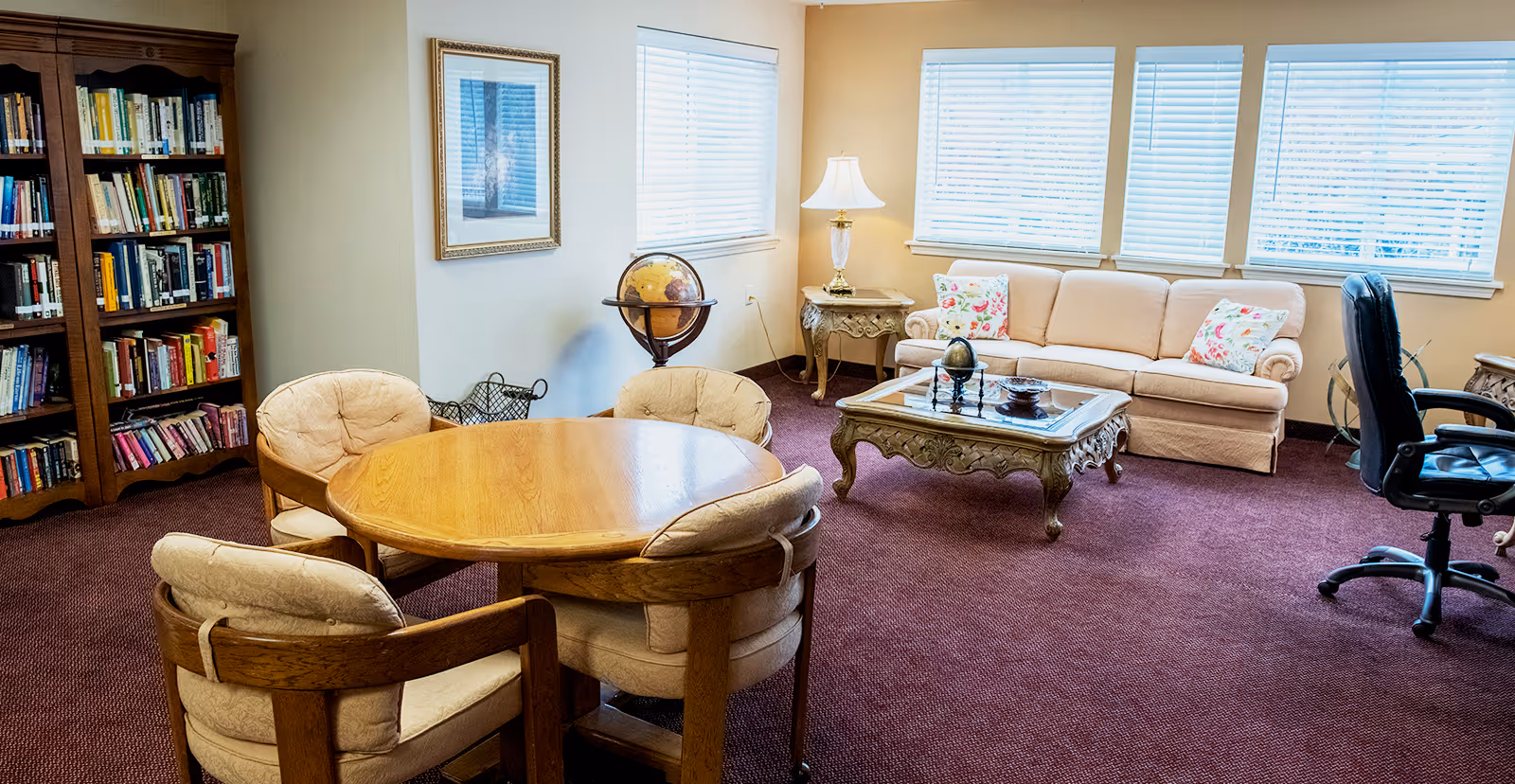 A cozy living room area with a wooden round table surrounded by four cushioned chairs, a beige sofa with floral pillows, a glass-top coffee table with decorative items, a wooden bookshelf filled with books, a globe on a stand, a side table with a lamp, and large windows with blinds letting in natural light.