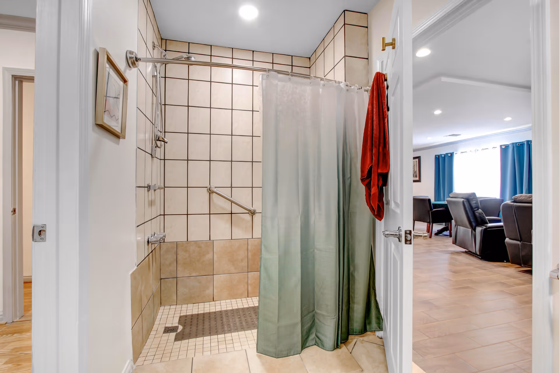 View of a tiled shower area with a green shower curtain partially drawn, a grab bar on the wall, and a red towel hanging on the door. The shower has a tiled floor and walls with beige and brown tiles. Through the open door, a living room area with dark chairs, a table, blue curtains, and a window letting in natural light is visible.