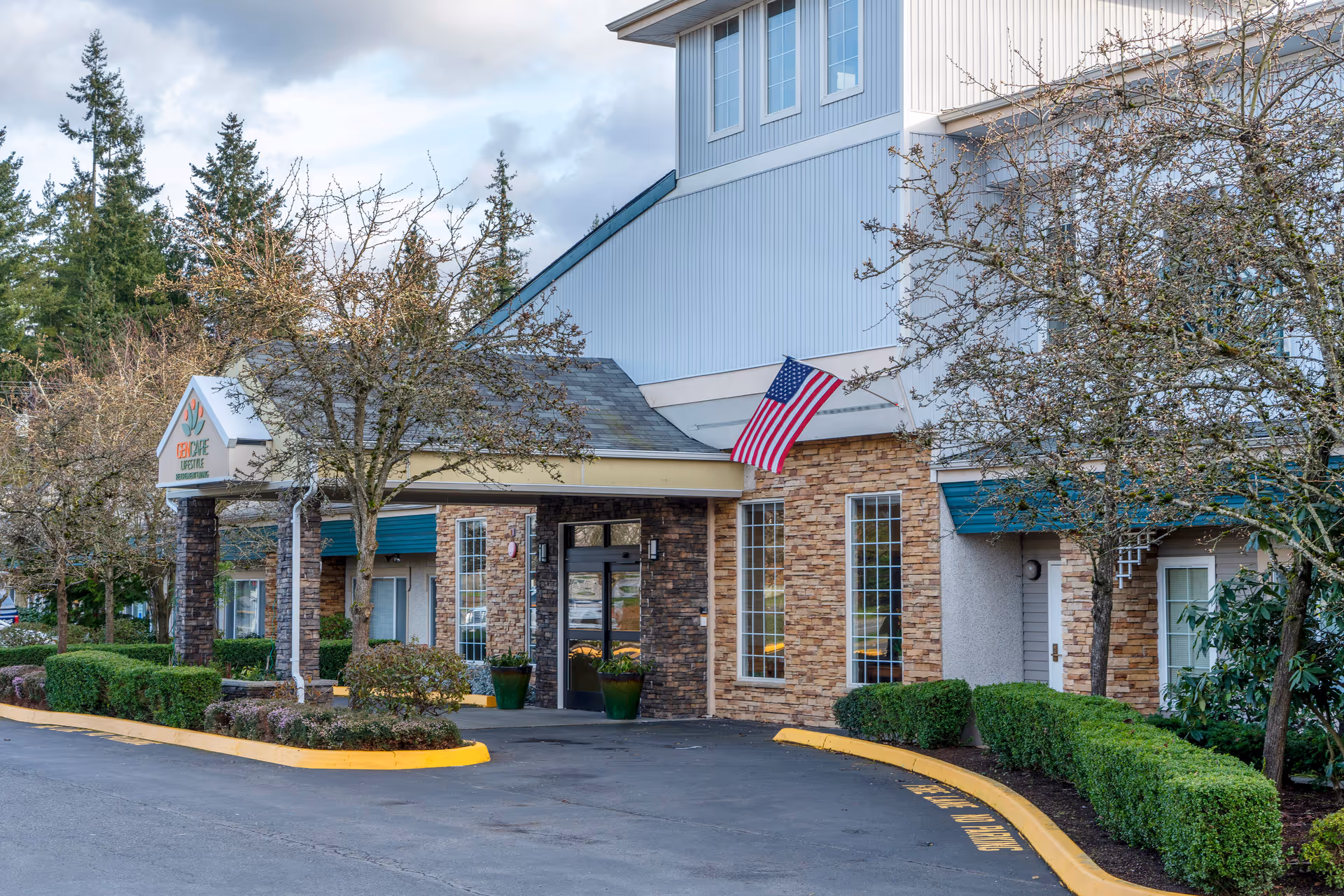 Exterior view of the entrance to GenCare Lifestyle Lynnwood at Scriber Gardens, featuring a covered driveway, stone and siding facade, an American flag, and landscaped bushes and trees.