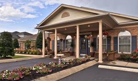 Front exterior view of a single-story brick building with a covered porch supported by columns, flower beds with colorful flowers, and a paved driveway under a partly cloudy sky.