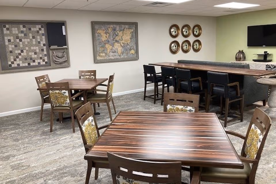 Interior view of a common area with multiple wooden tables and chairs. The chairs have floral patterned cushions. On the walls, there is a world map and a decorative piece with circular mirrors. A television is mounted on the wall in the background.