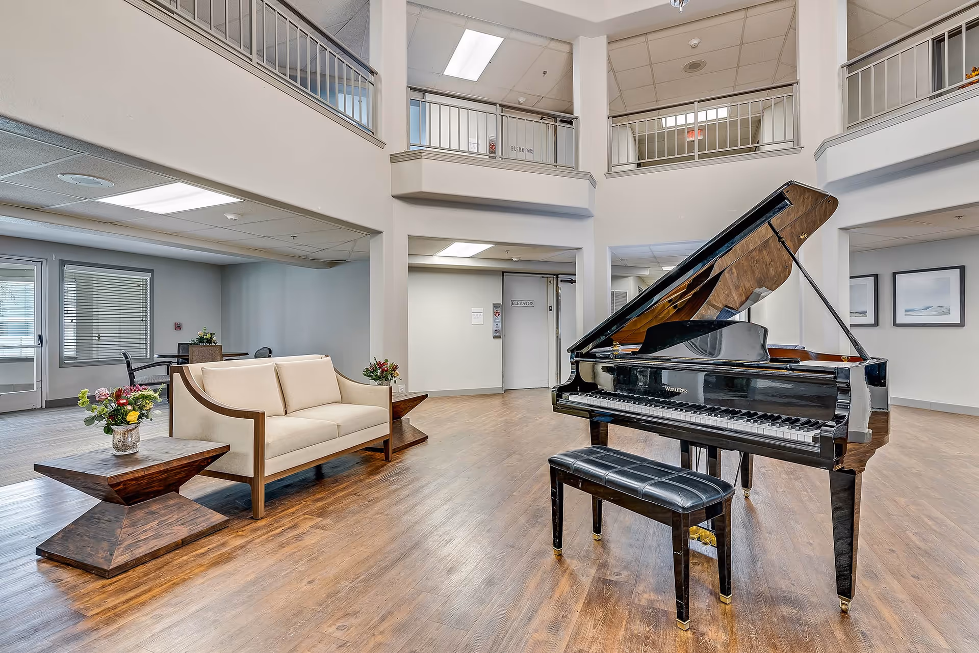 Spacious senior living common area with a black grand piano and bench, a sofa with side tables and floral arrangements, and a two-story open atrium.