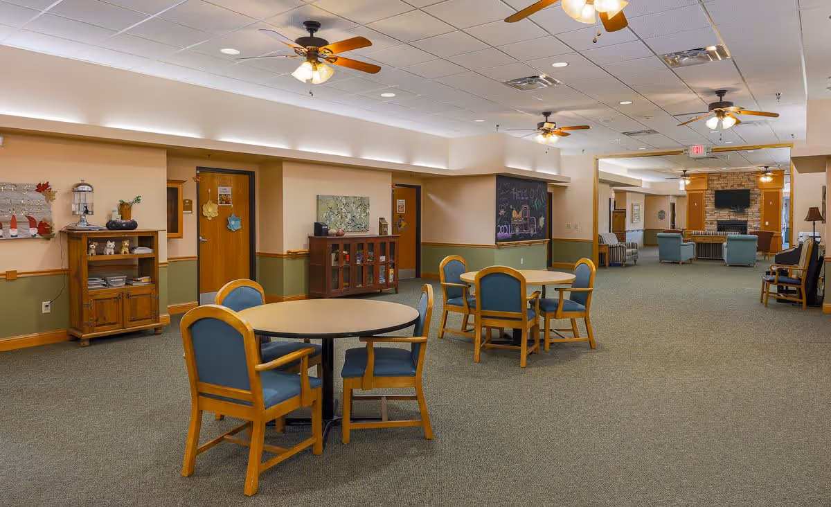 A spacious common area in an assisted living facility featuring round tables with blue cushioned chairs, ceiling fans with lights, a wooden cabinet with decorative items, a chalkboard on the wall, and a seating area with armchairs and a TV mounted above a stone fireplace in the background.