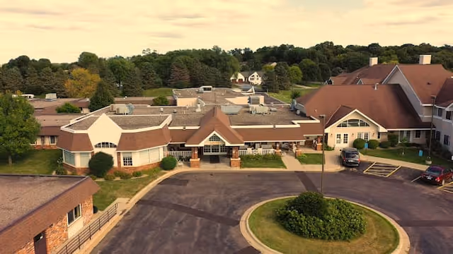Aerial view of Mala Strana Assisted Living facility showing multiple connected buildings with brown roofs, surrounded by greenery and trees. There is a circular driveway with a landscaped center island in front of the main entrance, and several parked cars are visible.