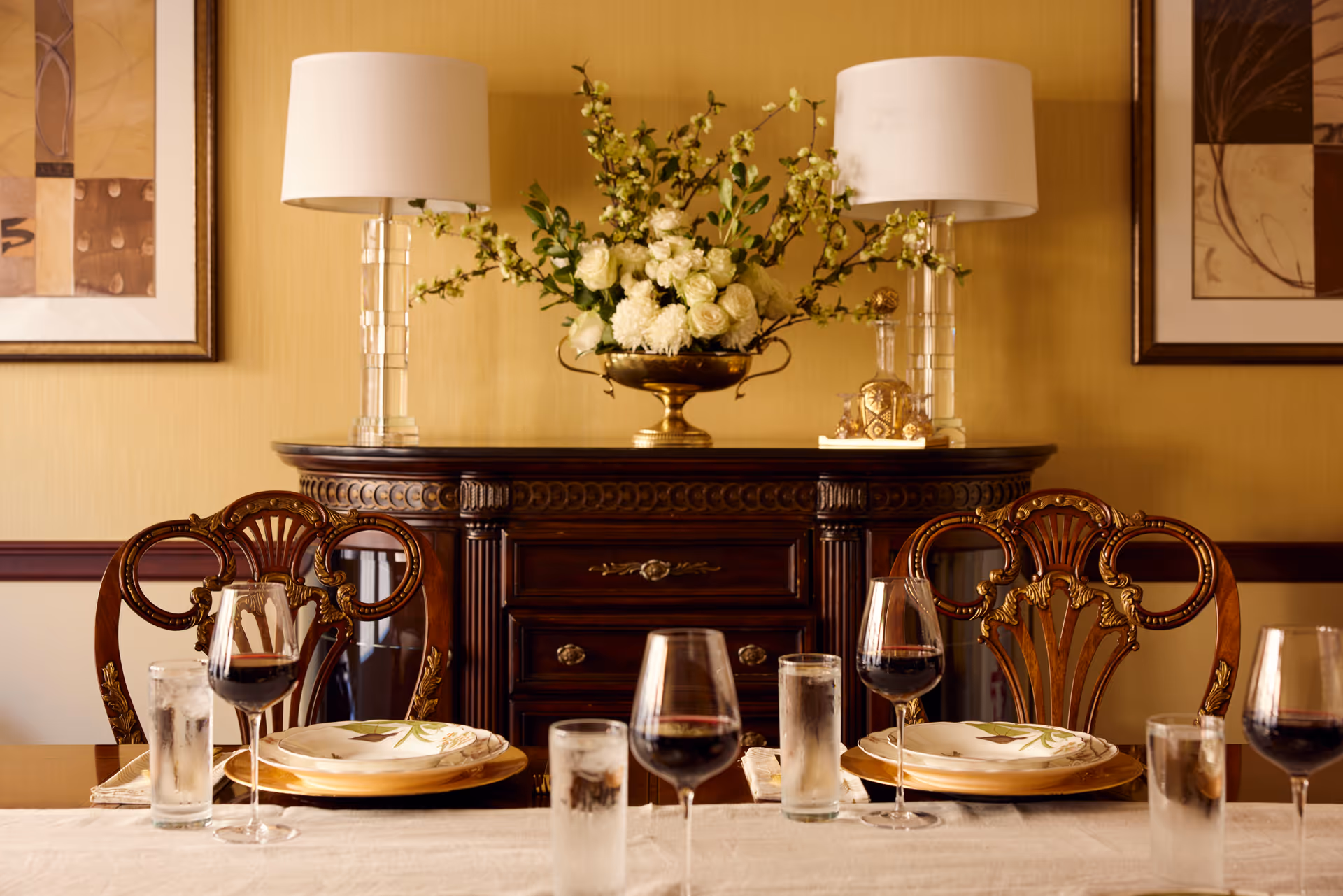 A formal dining room setup with a wooden table set for four, featuring plates, wine glasses filled with red wine, and water glasses. Behind the table is a dark wooden sideboard with decorative carvings, topped with two clear glass lamps with white shades and a large floral arrangement in a gold vase. The walls are painted yellow and adorned with framed artwork.