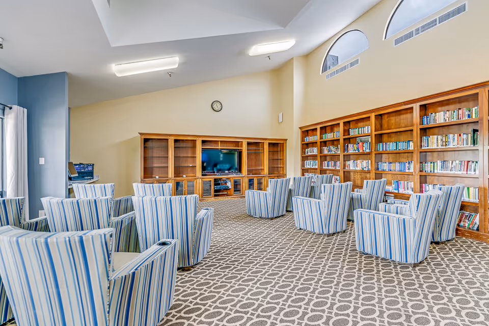 A spacious senior living common area with striped armchairs arranged in rows facing a wooden entertainment center with a television. The room features large wooden bookshelves filled with books, a patterned carpet, high ceilings with arched windows, and a wall clock above the entertainment center.