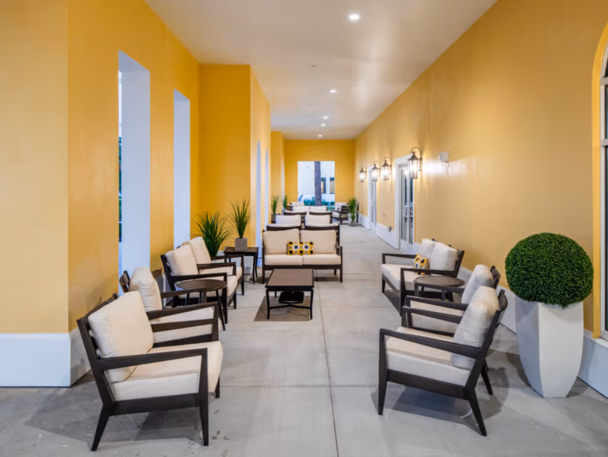 A long, covered outdoor seating area with yellow walls and multiple sets of cushioned chairs and sofas arranged around small tables. There are potted plants along the walls and wall-mounted lantern-style lights providing illumination.