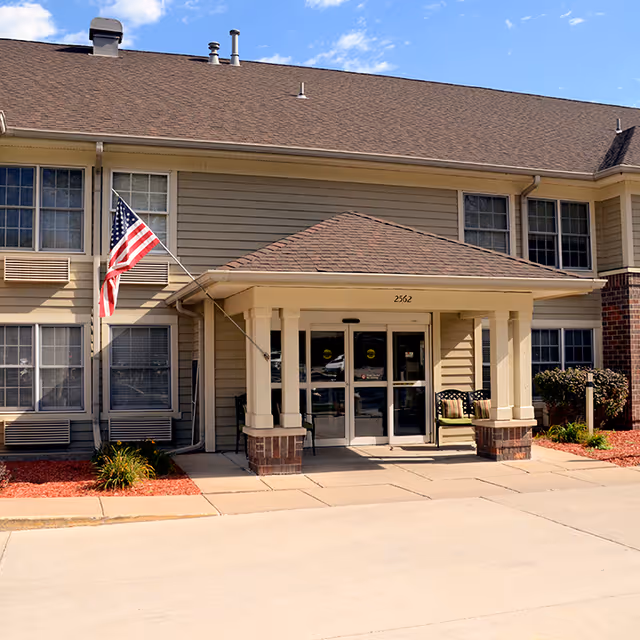 Front entrance of a senior living facility with a covered porch supported by columns, an American flag mounted on the left side, and two chairs with cushions on the right side. The building has beige siding, multiple windows, and a brown shingled roof under a partly cloudy sky.
