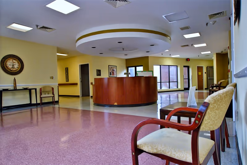 Interior view of a senior living facility reception area with a circular wooden front desk in the center, beige walls, and several chairs with patterned upholstery arranged along the walls. The floor has a pink and white tile pattern, and there are large windows letting in natural light.