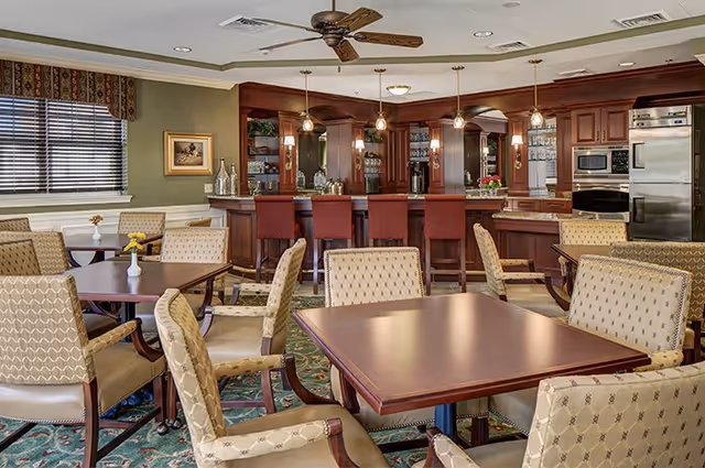 A spacious dining area with multiple tables and cushioned chairs arranged neatly. In the background, there is a wooden bar counter with high stools, pendant lights hanging above, and a kitchen area with stainless steel appliances including a refrigerator and oven. The room has a patterned carpet, a ceiling fan, and a window with blinds and a valance.