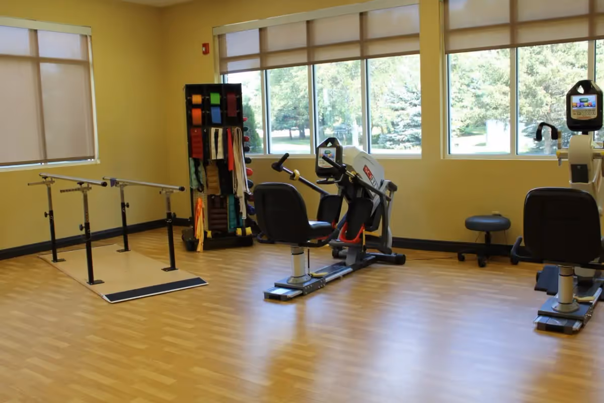 A rehabilitation or exercise room with parallel bars for walking support, two seated exercise machines, a stool, and a rack holding various resistance bands and exercise equipment. Large windows with beige blinds allow natural light and show trees outside.
