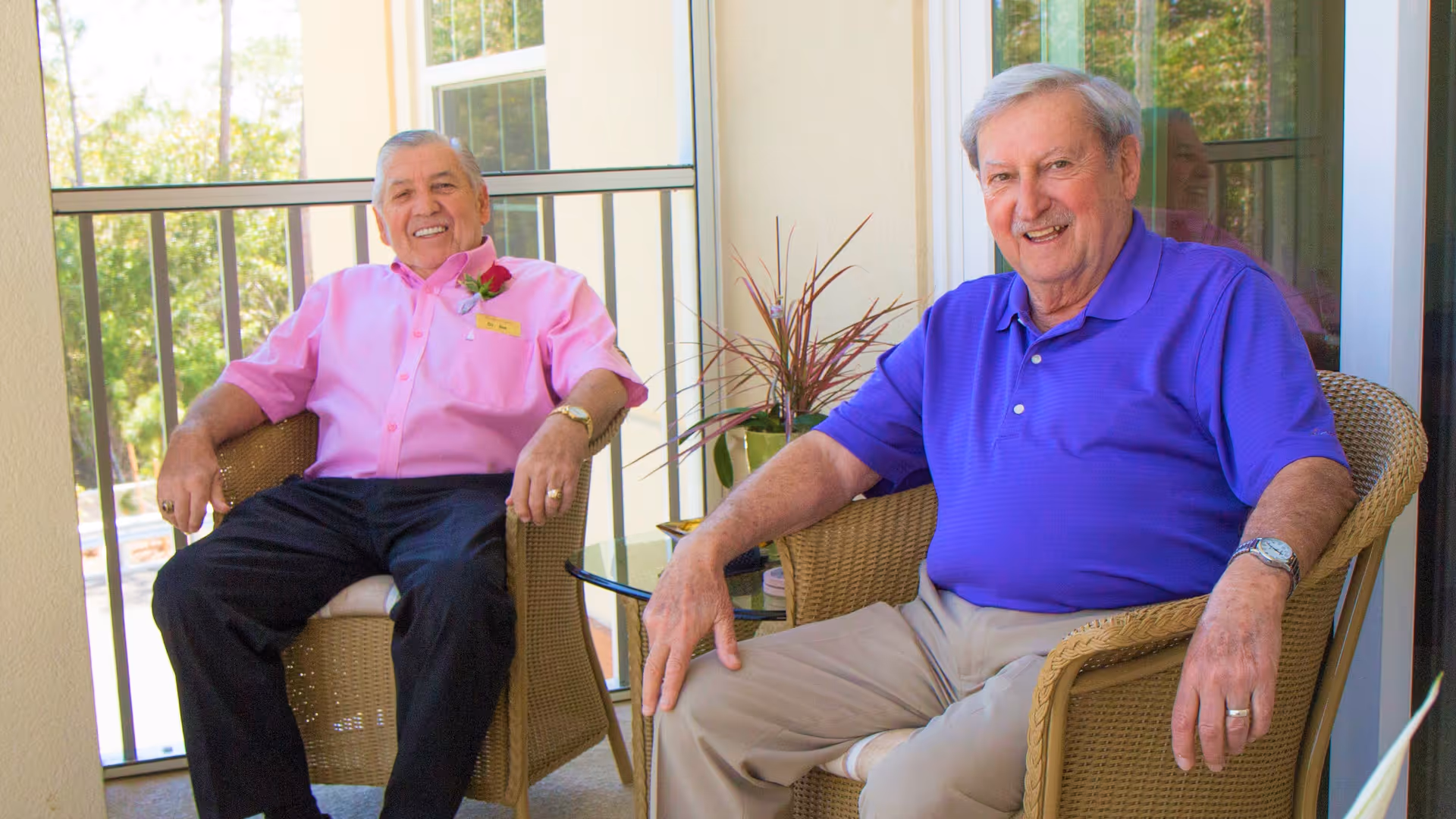 Two elderly men sitting and smiling on wicker chairs on a balcony or patio area with a small glass table and a potted plant between them.