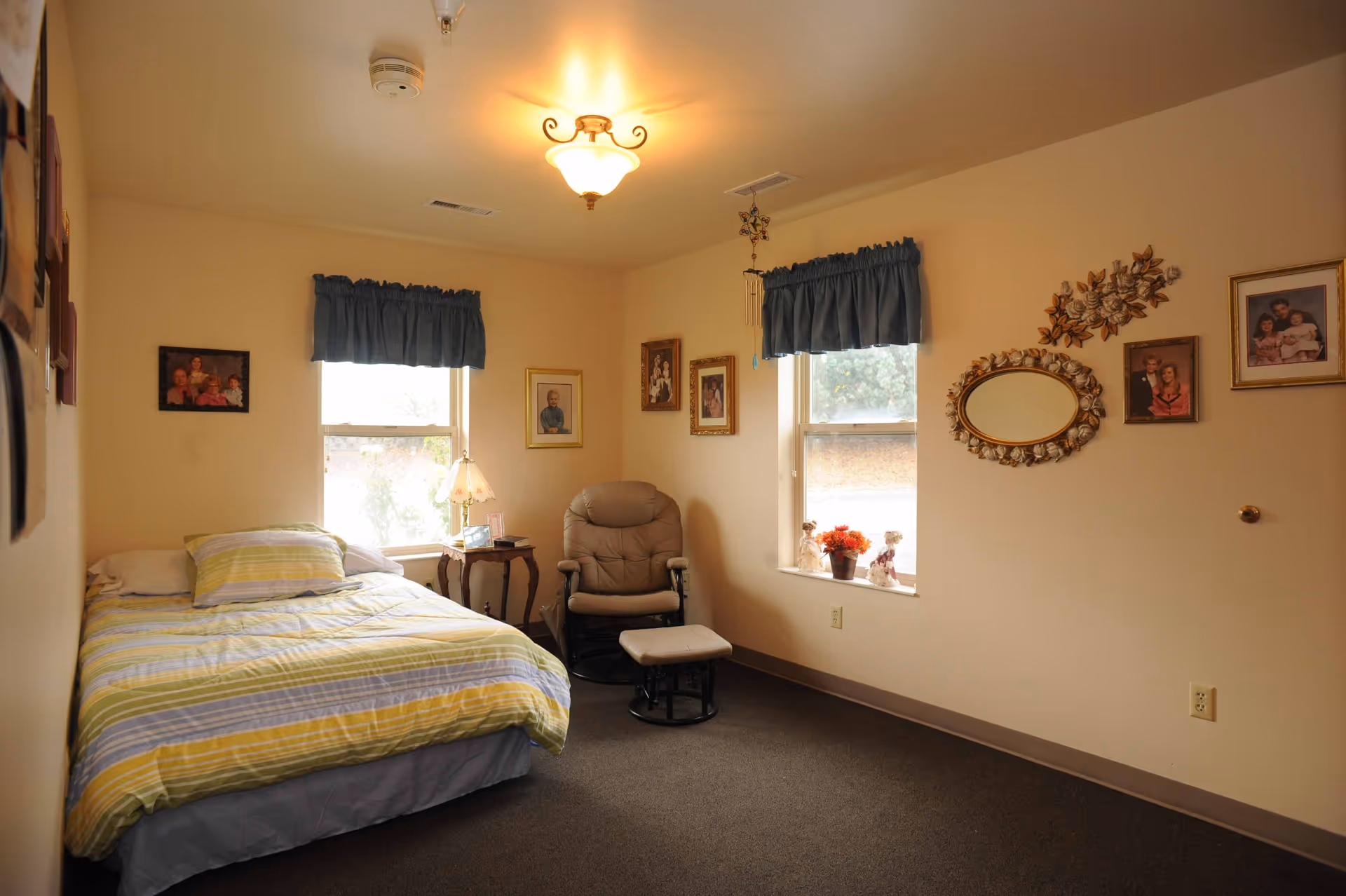 A cozy bedroom with a single bed covered in a striped yellow, gray, and white comforter. There are two windows with blue valances, a small wooden side table with a lamp, and a cushioned armchair with an ottoman. The walls are decorated with several framed family photos and a decorative oval mirror with a floral frame.