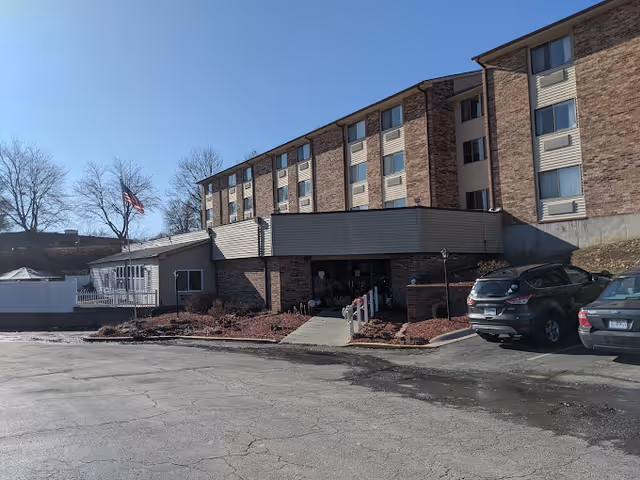 Exterior view of a multi-story brick building with several windows, a main entrance with a covered walkway, a few parked cars, and an American flag on a flagpole to the left. The sky is clear and blue.