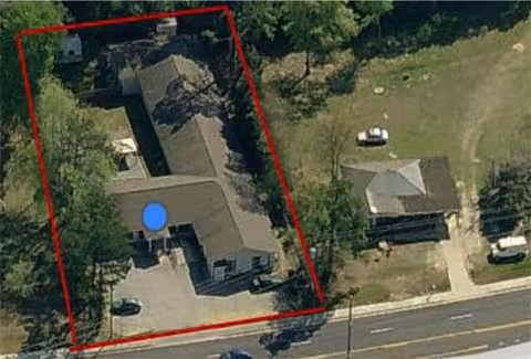 Aerial view of a property outlined in red, showing a building with a parking area and surrounding trees, adjacent to a road with vehicles and neighboring buildings.