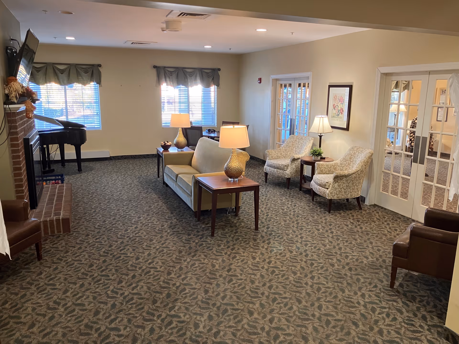 A cozy living room area in a senior living facility with a beige sofa, two patterned armchairs, two table lamps on wooden side tables, a fireplace with a mounted TV above it, and a grand piano near two windows with green valances. There are double glass doors on the right side leading to another room.