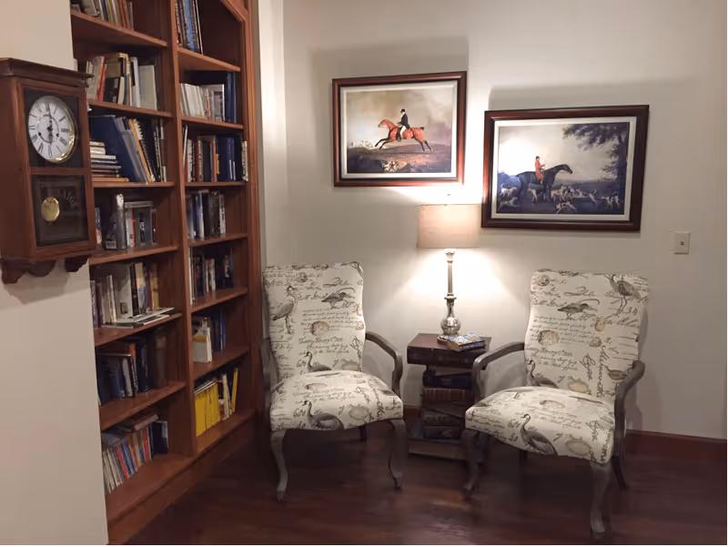 Two patterned armchairs flank a small table with a lamp in a cozy reading nook with bookshelves and framed equestrian paintings on the wall.