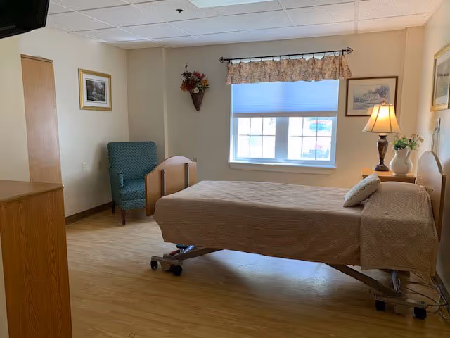 A simple, clean bedroom in a senior living facility with a single hospital-style bed covered in a beige quilt. There is a wooden nightstand with a lamp and a vase of flowers next to the bed. A green upholstered armchair is placed near the corner, and framed pictures hang on the light-colored walls. A window with a floral valance and a closed blind lets in natural light.