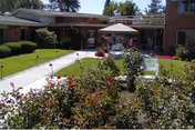 Outdoor garden area at Rockwood at Whitworth with a paved walkway, green lawn, blooming flowers, and a patio umbrella with seating in front of a single-story building.