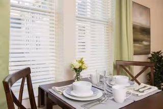 A dining table set for two with white bowls, plates, cups, and silverware. A small vase with yellow flowers is placed in the center of the table. The table is positioned in front of two large windows with white blinds and light green curtains. A framed artwork and a potted plant are visible in the background.