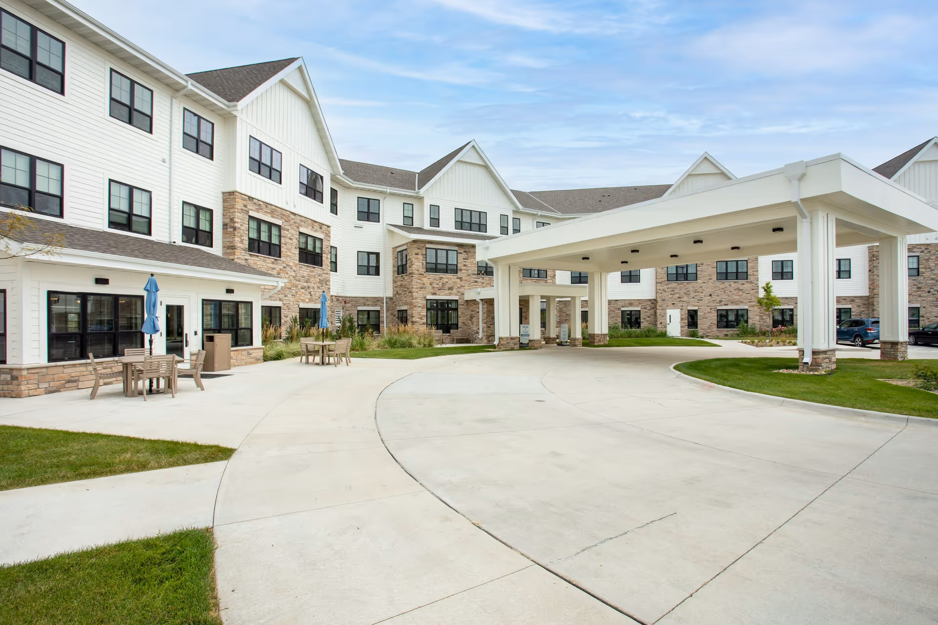 Exterior front view of a multi-story senior living building with a covered porte-cochere, paved driveway, and outdoor seating area.
