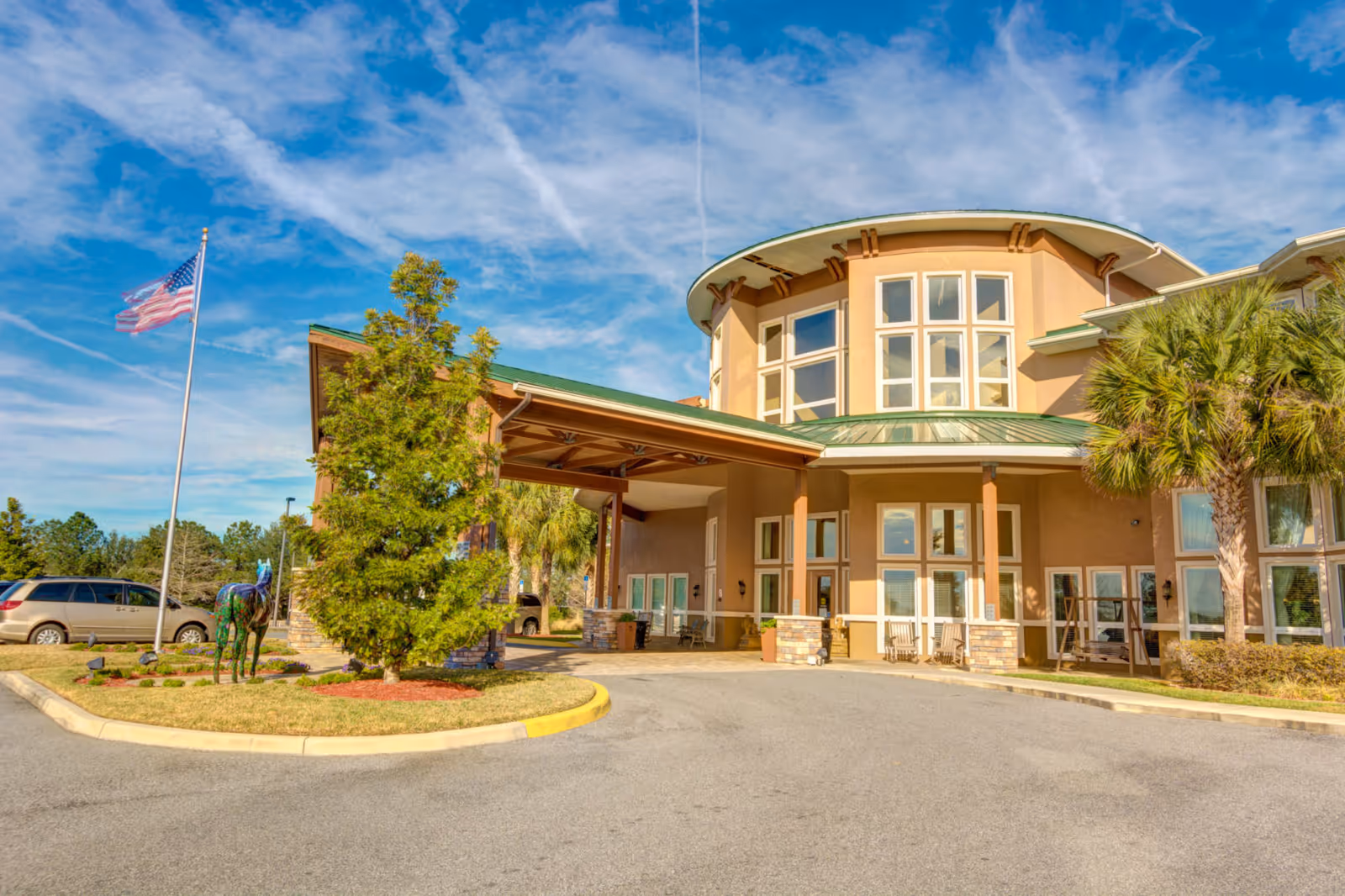 Exterior view of Brentwood at Fore Ranch facility showing a large building with a covered entrance, multiple windows, palm trees, an American flag on a flagpole, and a decorative horse sculpture on a landscaped island.