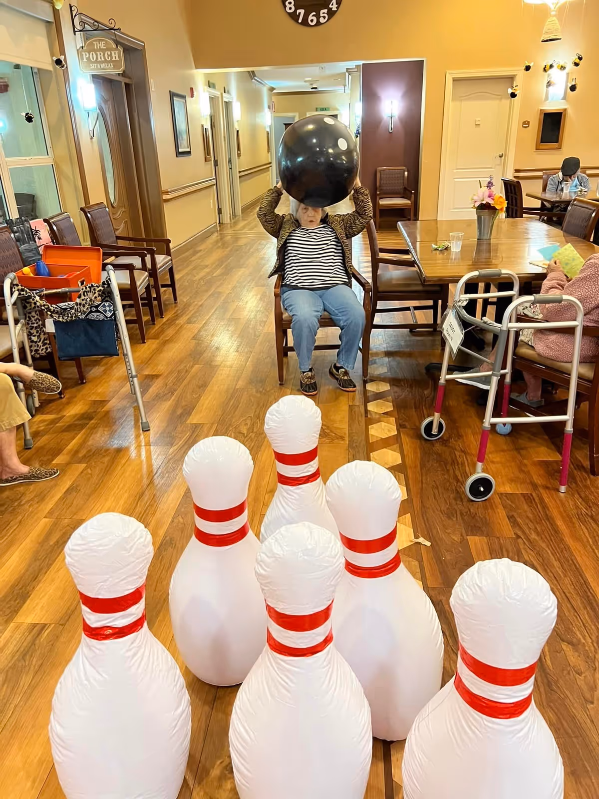 An elderly woman sitting on a chair in a hallway holding a large black bowling ball above her head, preparing to roll it towards six large inflatable bowling pins set up on the wooden floor. Other elderly individuals are seated around a table and along the hallway, some with walkers nearby. The setting appears to be a communal area in a senior living facility.