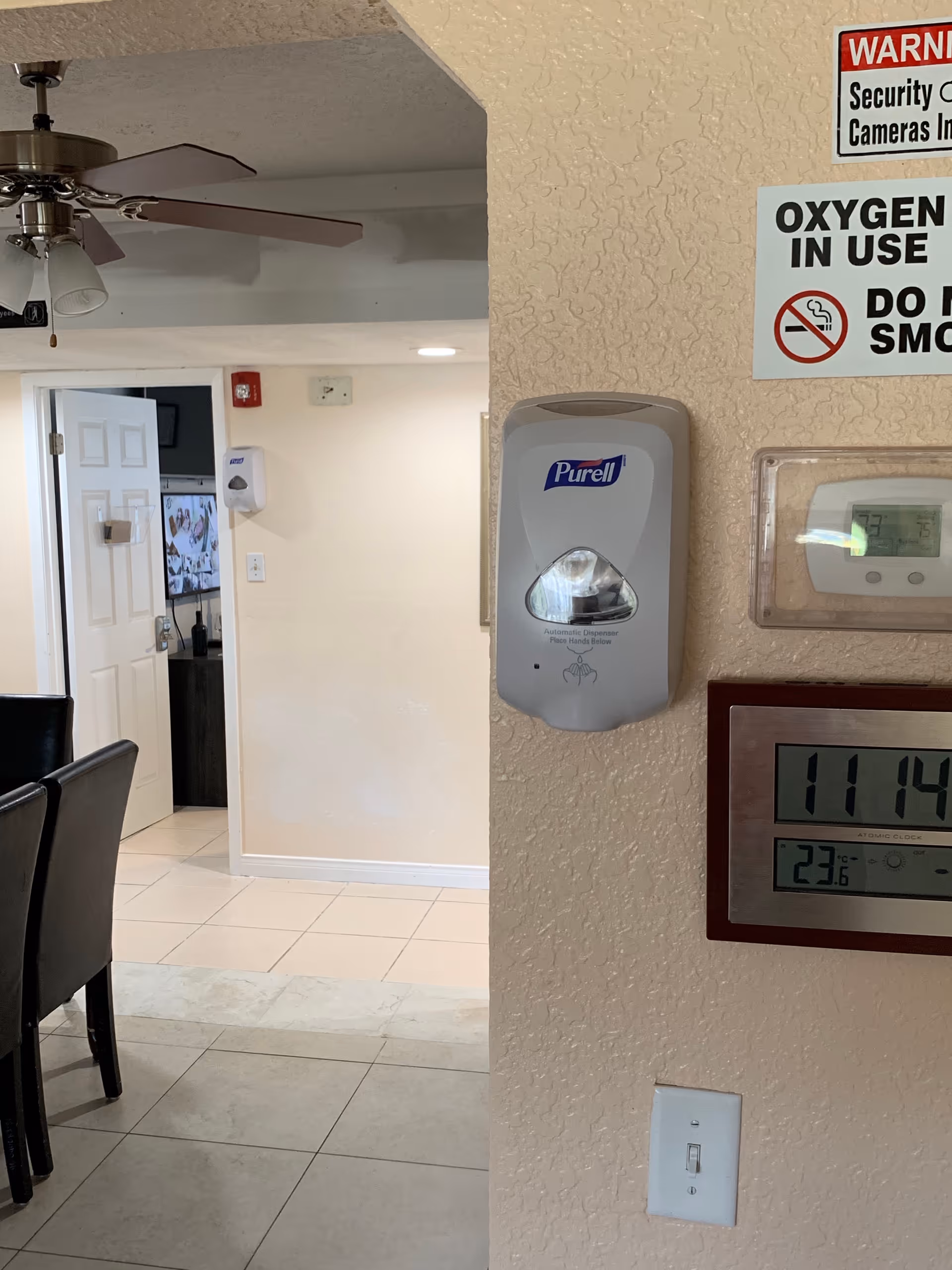Interior view of a room with a wall-mounted Purell hand sanitizer dispenser, a digital clock and thermostat, and warning signs about security cameras and no smoking due to oxygen use. In the background, there is an open door leading to another room with a television visible, a ceiling fan with lights, and part of a dining table with chairs.