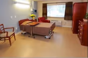 Private patient room with an adjustable hospital bed, overbed table, dresser, chairs, and a window.