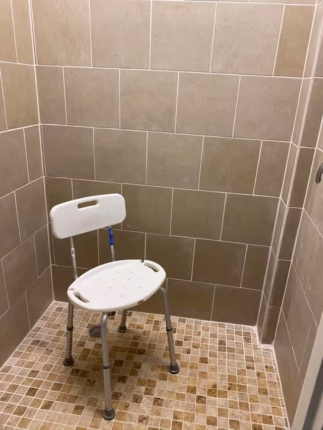 A shower area with beige tiled walls and a mosaic tiled floor, featuring a white plastic shower chair with metal legs and a backrest.