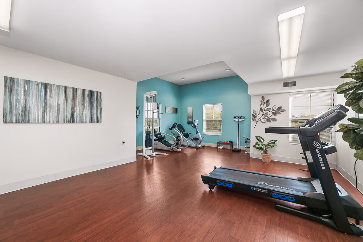 A bright fitness room with wooden flooring, featuring a treadmill, exercise bikes, a weight machine, and a scale. The walls are painted white and teal, decorated with abstract artwork and a metal leaf wall sculpture. There are two windows letting in natural light and some potted plants adding greenery to the space.
