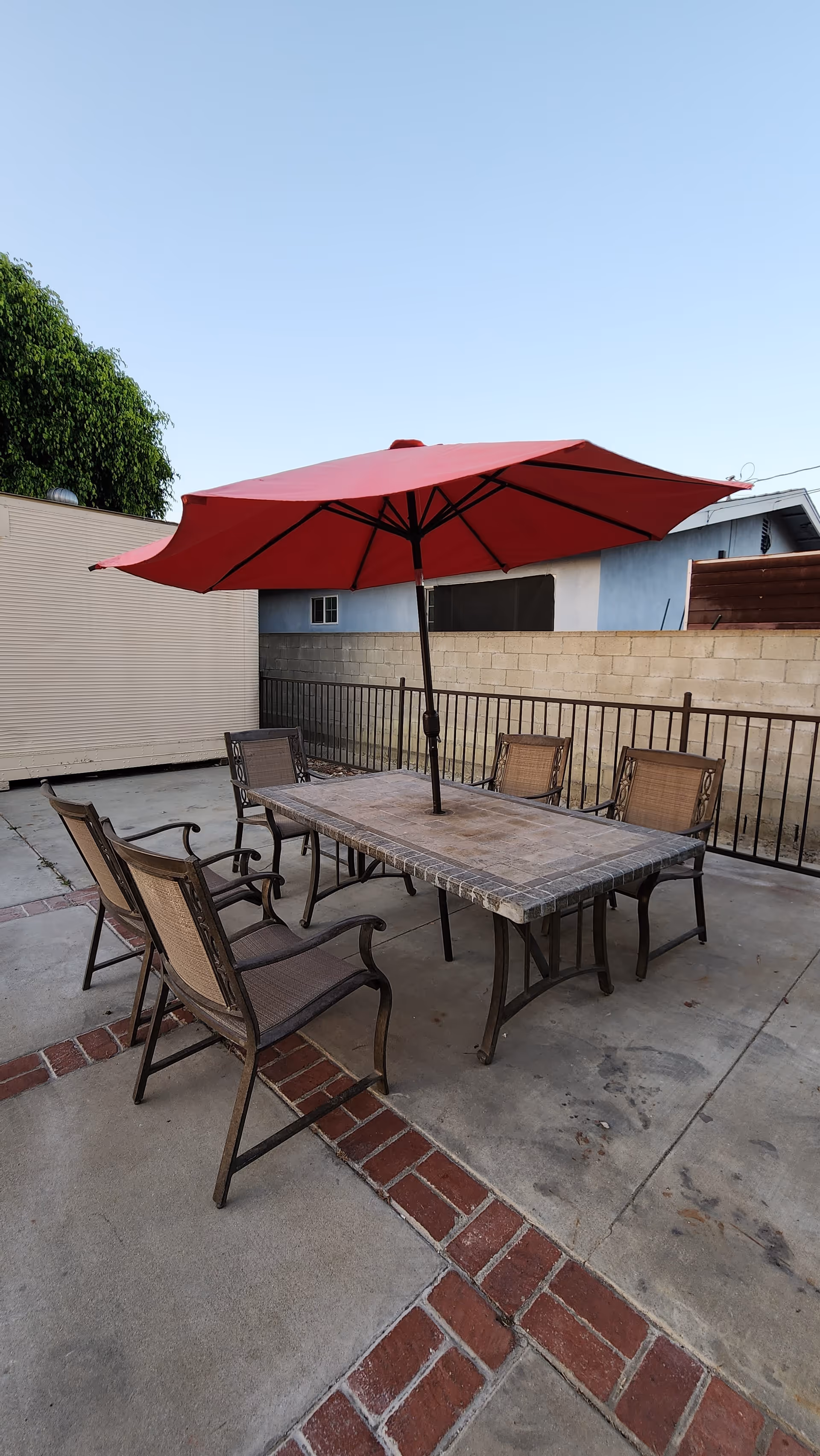 Outdoor patio area with a rectangular stone table surrounded by six metal and mesh chairs. A large red umbrella is positioned in the center of the table providing shade. The patio is paved with concrete and bordered with red bricks. There is a metal fence and a beige brick wall in the background, along with parts of neighboring buildings and a tree visible.