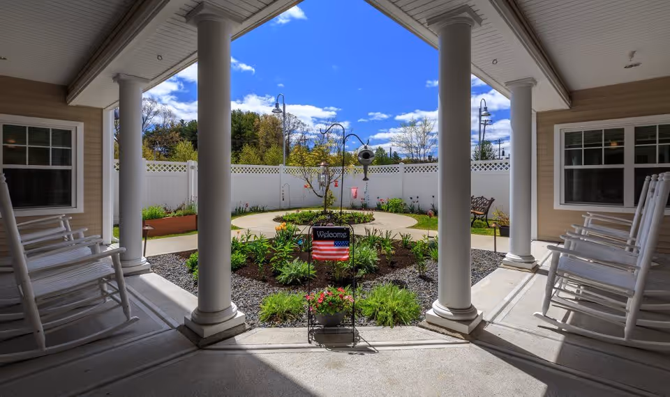 Covered outdoor patio area with white rocking chairs on both sides, white columns, and a landscaped garden with a circular walkway and a small welcome sign featuring an American flag in the center. The area is enclosed by a white fence with trees and a blue sky with clouds in the background.