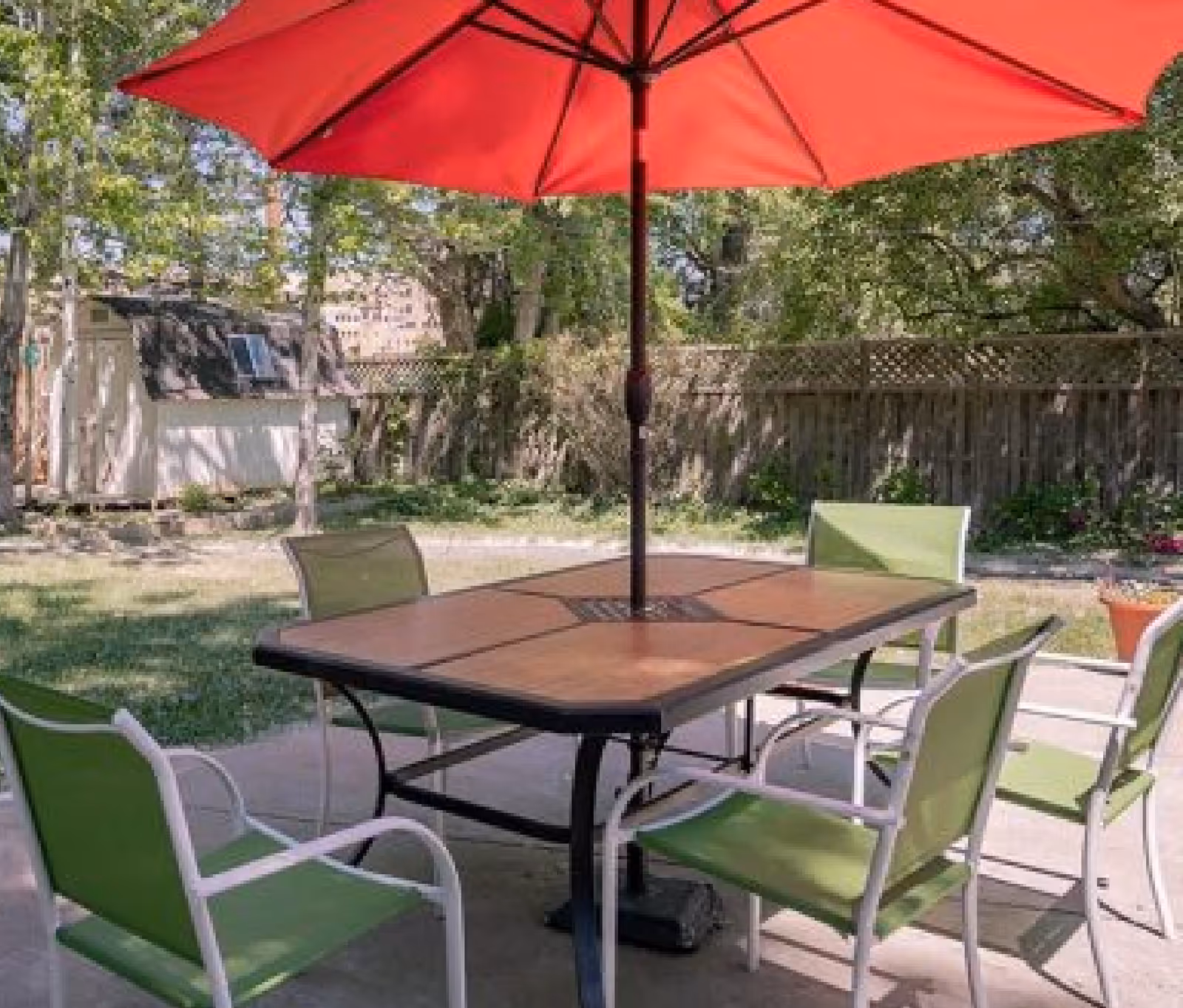 Outdoor patio area with a rectangular wooden table surrounded by six green and white chairs. A large red umbrella is positioned in the center of the table providing shade. In the background, there is a wooden fence, trees, and a small white shed.
