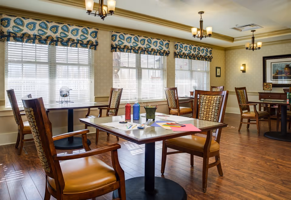 A bright and inviting room with multiple wooden tables and chairs arranged neatly. The tables have various art supplies including colored paper, markers, and paint bottles. Large windows with patterned valances allow natural light to fill the space. The room has warm wooden flooring, soft yellow walls, and ceiling light fixtures.