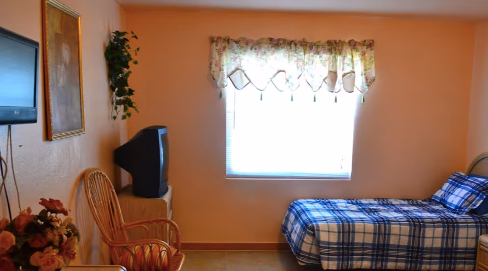 A small bedroom with peach-colored walls, a single bed with blue and white plaid bedding, a window with floral valance curtains, a wooden chair, a small TV on a stand, a wall-mounted flat screen TV, a framed picture, and a vase with flowers.
