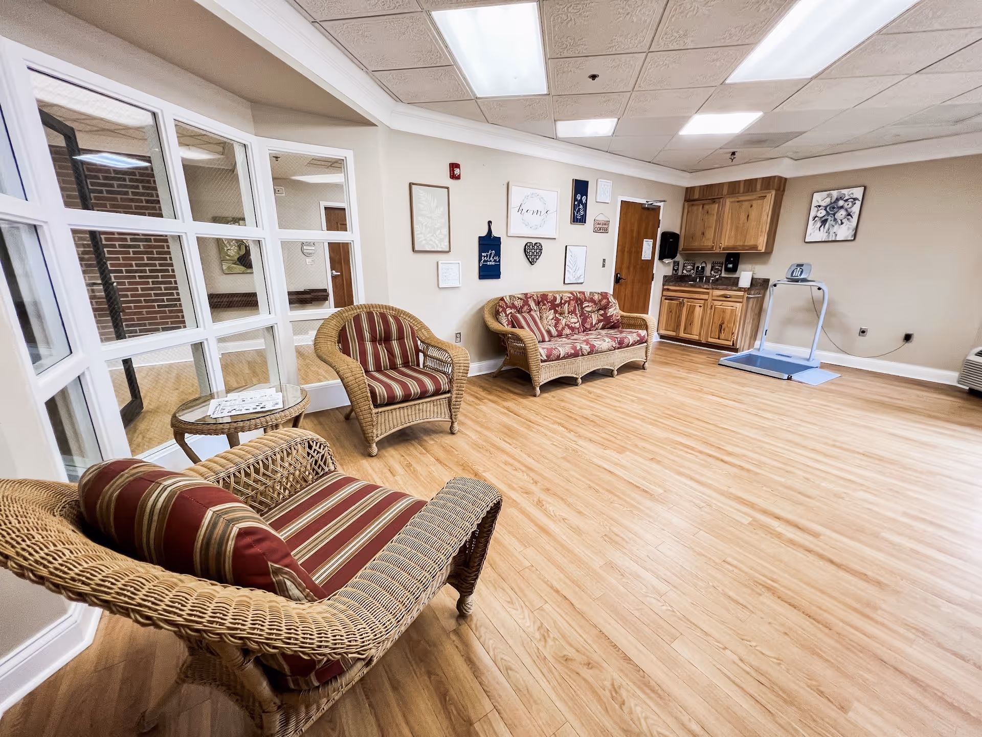 A cozy sitting area with wicker furniture including two armchairs and a loveseat with red and beige striped cushions. The room has light wood flooring, a small round wicker table with papers on it, and a wall decorated with framed art and signs. There is a wooden cabinet with a coffee station and a medical scale on a blue mat in the corner. The ceiling has white tiles with fluorescent lighting, and a large window with white framing looks into another room.