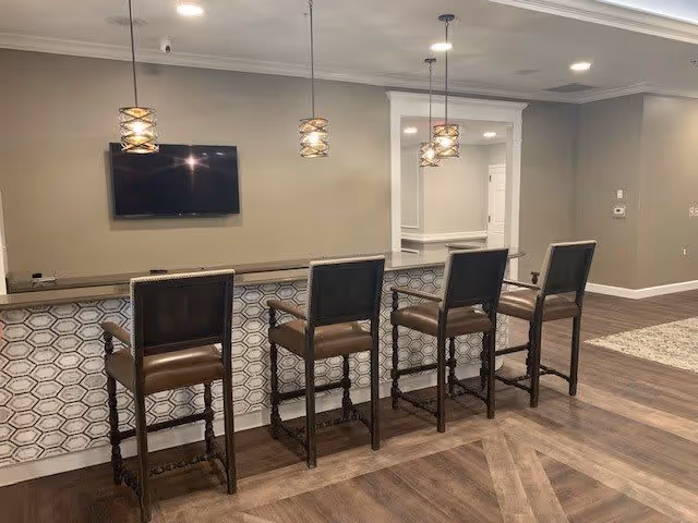 Interior view of a seating area with four high-back bar stools lined up against a counter with a geometric tile front. Above the counter hang three pendant lights with decorative shades. A flat-screen TV is mounted on the beige wall behind the counter. The floor is wood with a patterned inlay, and there is an open doorway leading to another room.