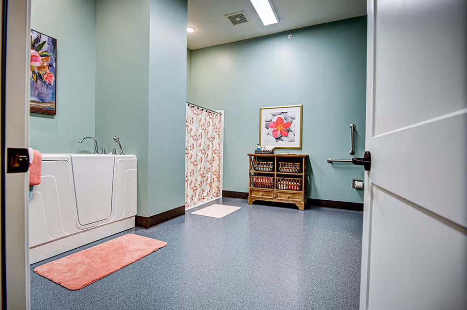 A spacious bathroom with light green walls and blue flooring. The room features a white walk-in bathtub with a pink bath mat in front, a floral shower curtain, a wooden cabinet with towels, a framed flower picture on the wall, and a grab bar next to the toilet paper holder.