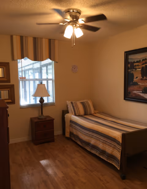 A small bedroom with a single bed covered in a striped bedspread in shades of beige, brown, and blue. There is a wooden nightstand with a lamp next to the bed, a window with blinds and a striped valance, two framed mirrors on the wall, a ceiling fan with lights, and a framed landscape painting on the wall.