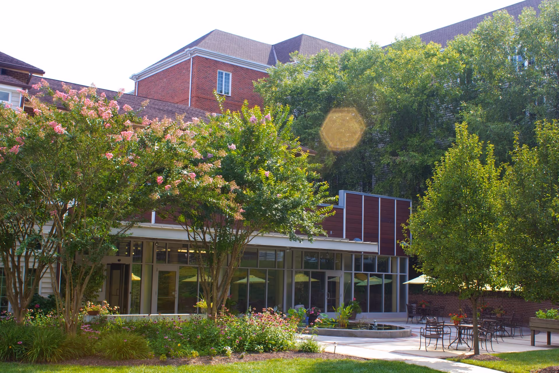 Outdoor courtyard area at Heart Lands Senior Living Village At Ellicott City featuring green trees, flowering plants, patio tables with chairs and umbrellas, and a building with large windows in the background.