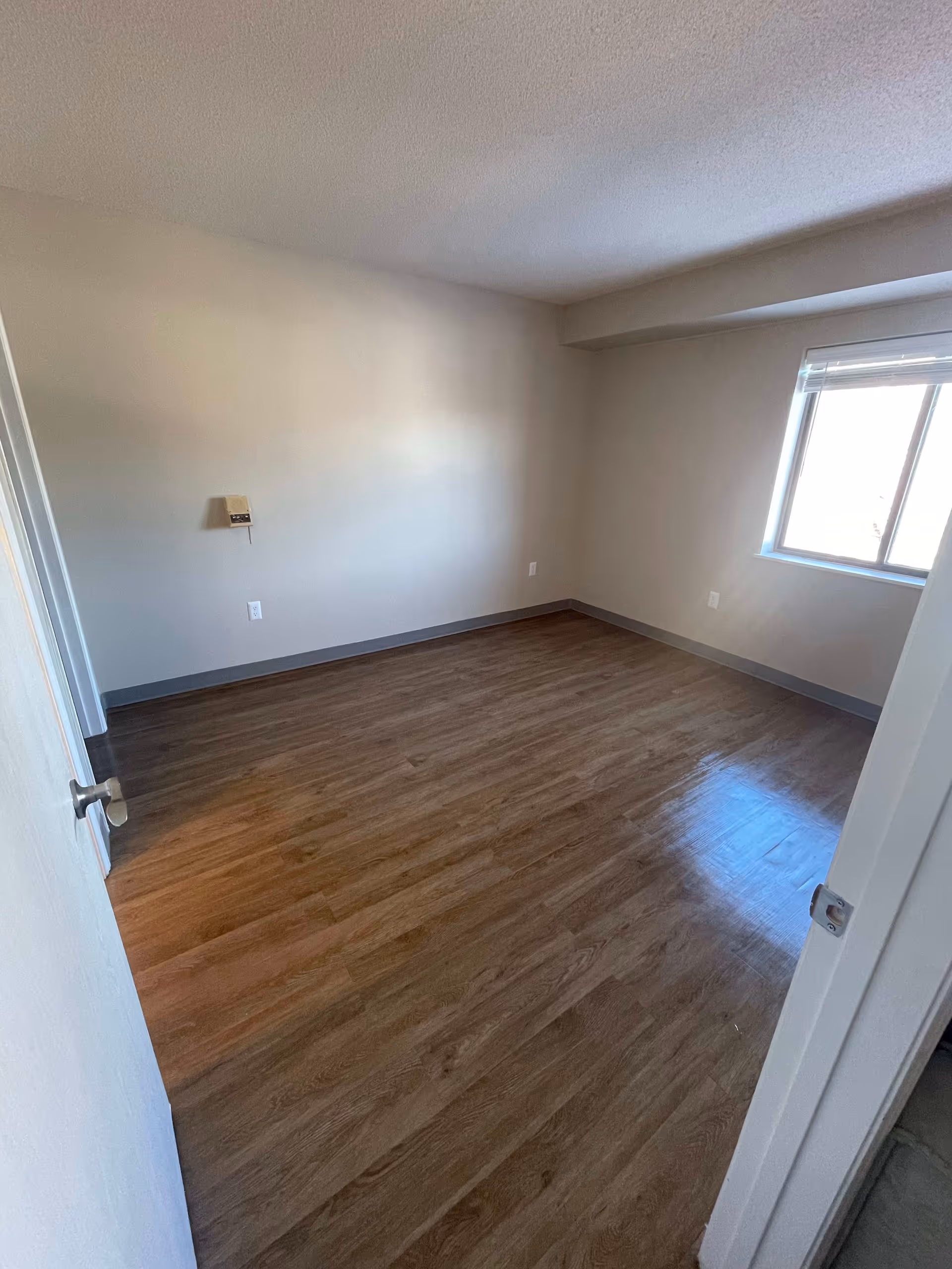 Empty room with wooden flooring, beige walls, a window with blinds letting in natural light, and a partially open door.