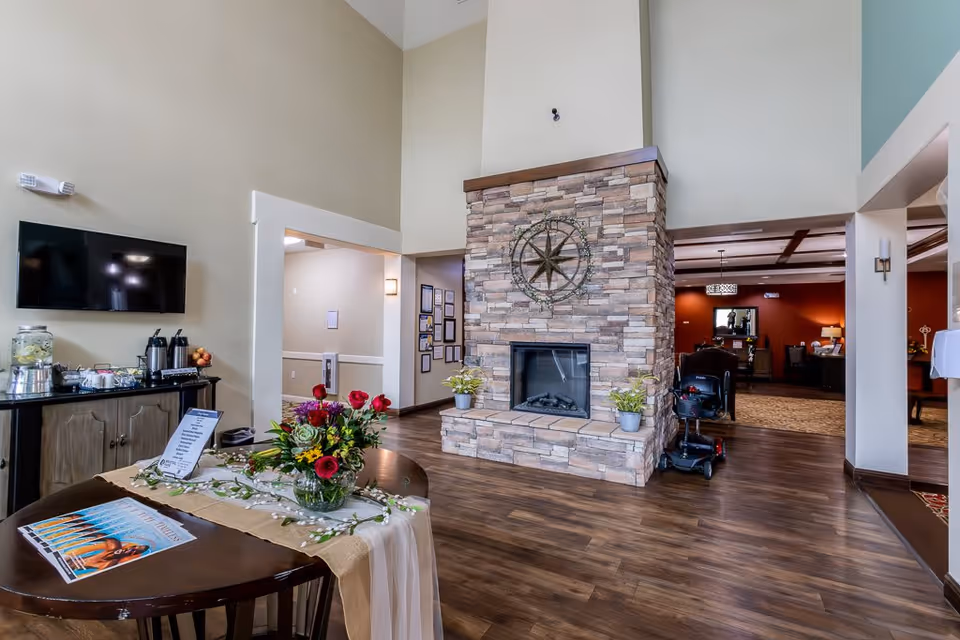 A spacious living area with a high ceiling featuring a stone fireplace decorated with a large metal compass rose. In front of the fireplace is a wooden table with a floral arrangement and magazines. To the left, a cabinet holds a beverage dispenser and coffee pots beneath a mounted flat-screen TV. The room has wood flooring and leads into another room with warm lighting and furniture.
