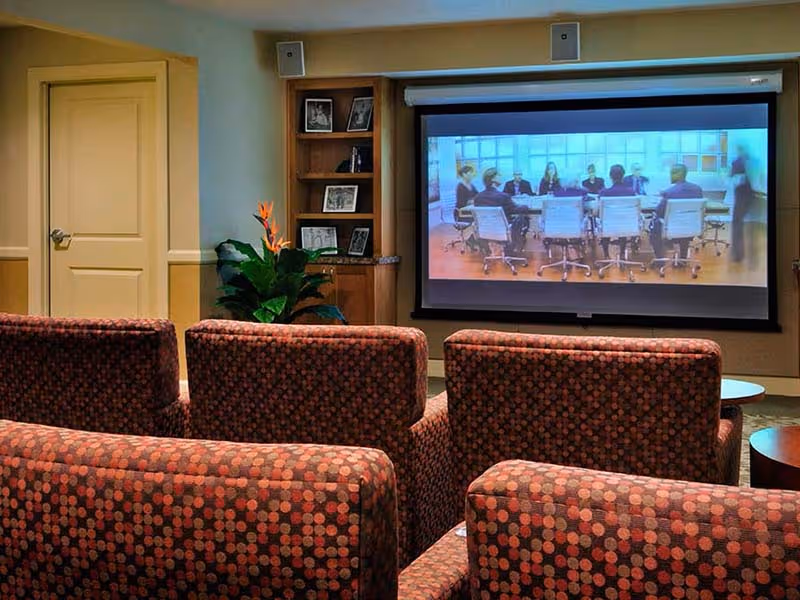 A cozy room with patterned upholstered chairs arranged in rows facing a large projection screen displaying a meeting scene. There is a wooden bookshelf with framed photos and a potted plant next to a closed door.