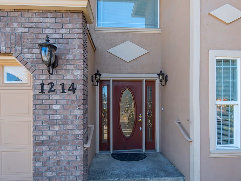 Front entrance of a building with a brown door featuring an oval glass panel and sidelights, flanked by two black wall-mounted lanterns. The building has a brick wall section with the number 1214 displayed and a beige stucco exterior with a window to the right.