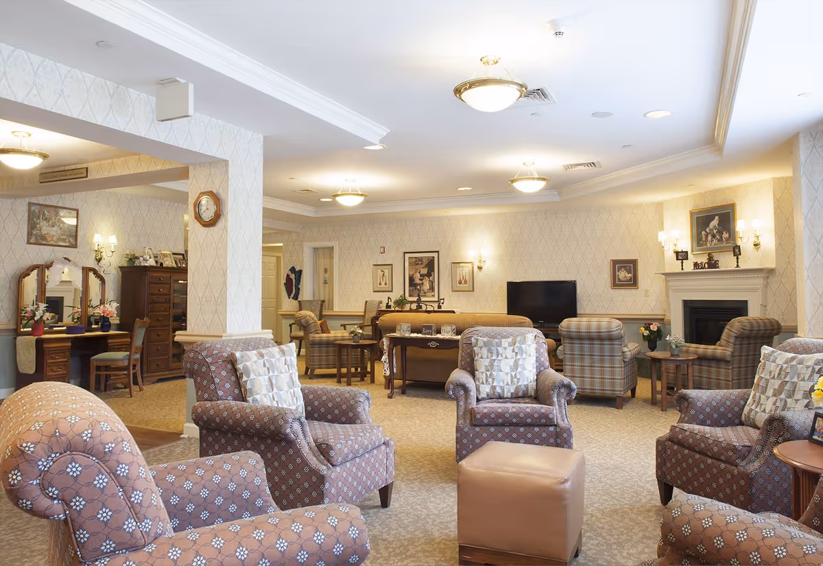 A cozy senior living facility common area with several upholstered armchairs arranged around a small ottoman. The room features patterned wallpaper, a fireplace with framed artwork above it, a flat-screen TV, and various tables with floral decorations. There is also a vanity area with a mirror and chairs visible in the background.