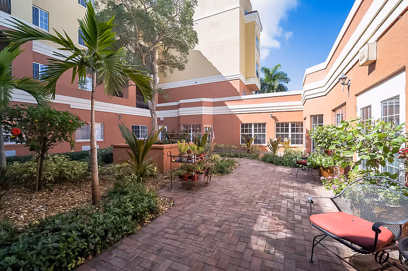 Walled sunlit courtyard with a brick paver path, tropical plants, and outdoor seating.